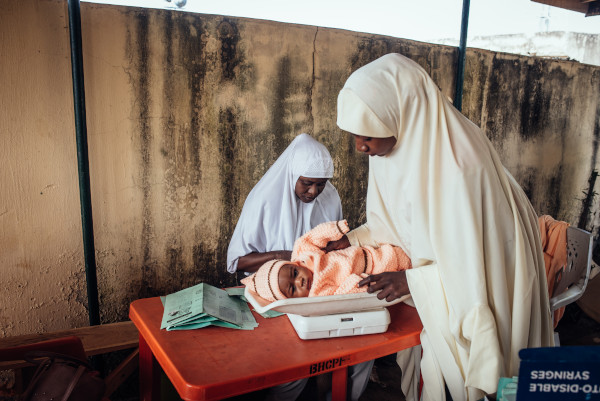 A mother and child wait at a clinic in Jigawa while a Routine Immunization Provider prepares paperwork before administering the infant's routine childhood immunizations.